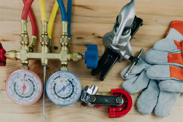 HVAC tools on a wooden table, including pressure gauges with colored hoses, pipe cutter, fittings, and a pair of work gloves.