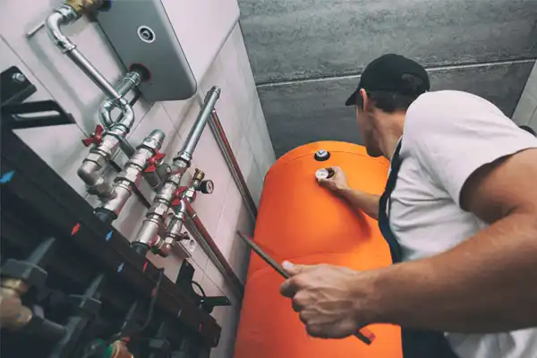 A technician adjusts a valve on an orange tank in a mechanical room, surrounded by pipes and gauges.
