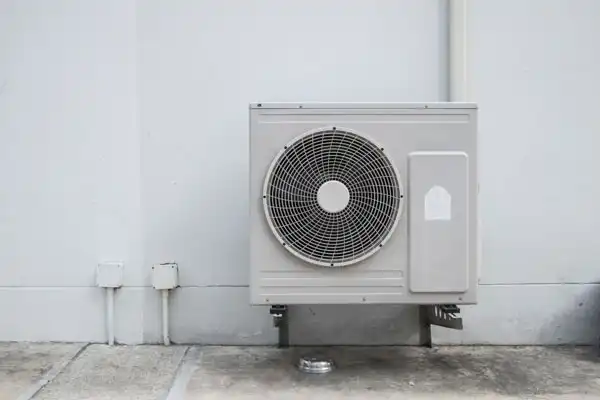 An outdoor air conditioning unit mounted on a wall with visible piping and a fan.