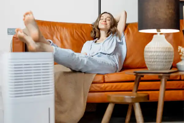 A person relaxes on a brown leather sofa, feet up, next to a dehumidifier and a lamp on a side table.