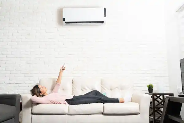 Woman lying on a sofa, holding a remote control, with a wall-mounted air conditioner above her against a white brick wall.
