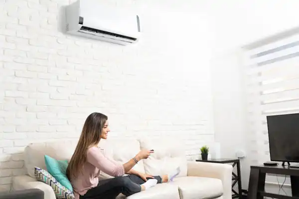 A person sits on a white sofa holding a remote control in a room with a wall-mounted air conditioner and TV.