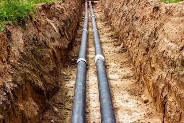 Two large black pipes run parallel in a trench dug into the ground, surrounded by dirt and grass.