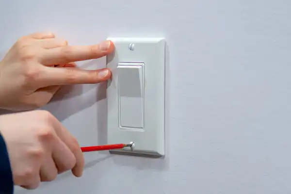 Person uses a red screwdriver to tighten a screw on a white light switch cover while steadying it with the other hand.