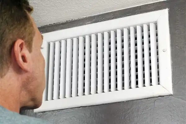 A person looks at a white vent cover on a gray wall.