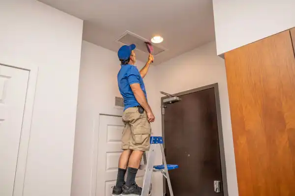 A man in a blue shirt and shorts stands on a ladder cleaning a ceiling air vent with a duster inside a room.