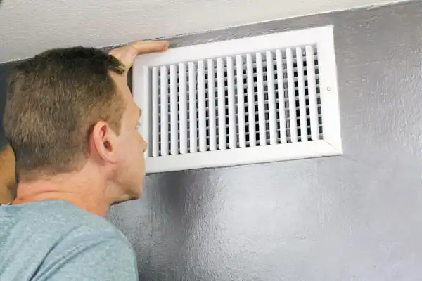 A person inspects a wall-mounted air vent with a white grille.