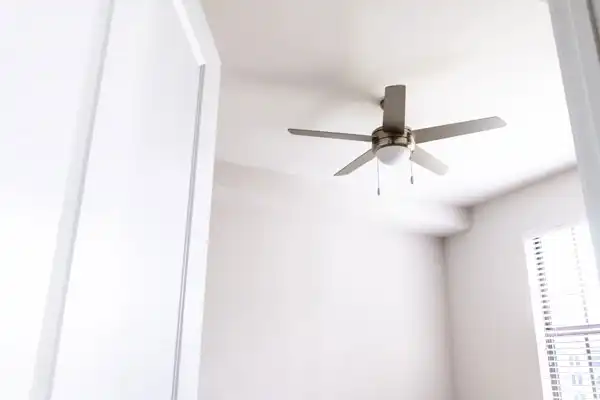 White ceiling fan with light fixture in a bright, minimalistic room. White walls and a window with blinds are visible.