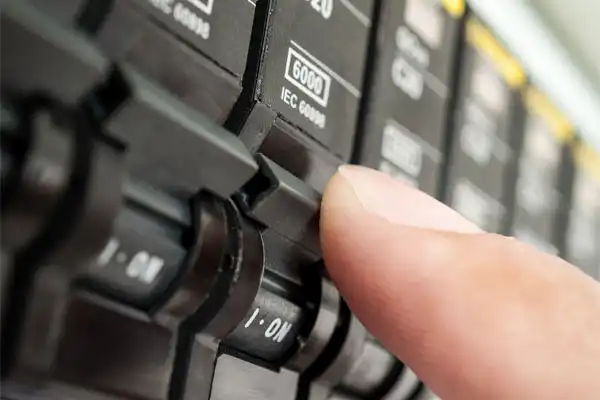 Close-up of a person's finger flipping a switch on an electrical circuit breaker panel with labels reading "6000" and "IEC 60906.