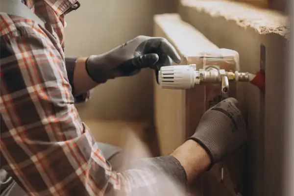 A person in a plaid shirt and gloves is adjusting a valve on a radiator.