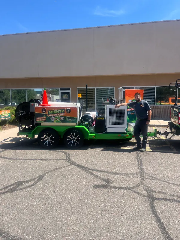 A person stands beside a green trailer-mounted pressure washer parked in front of a building with large windows.