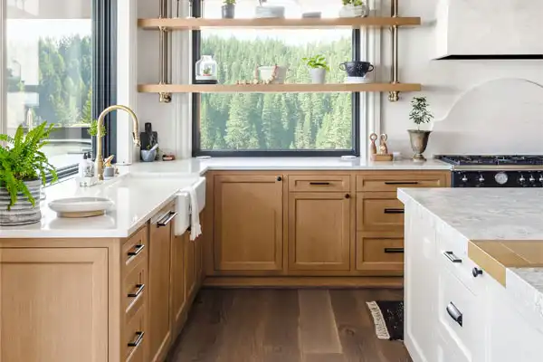 Modern kitchen with light wood cabinets, white countertops, and a large window with forest view. Open shelves with plants and dishes. Black stove and a sink with a gold faucet.