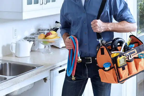 Person holding a plumbing tool bag and colorful hoses in a kitchen setting.