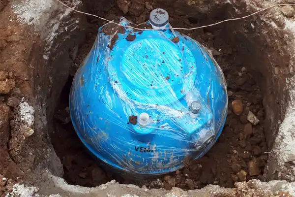 A blue water tank partially buried in a dirt hole, wrapped in protective plastic.
