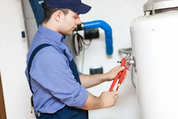 A plumber in a blue shirt and cap is using a red wrench to adjust a pipe on a white water heater.