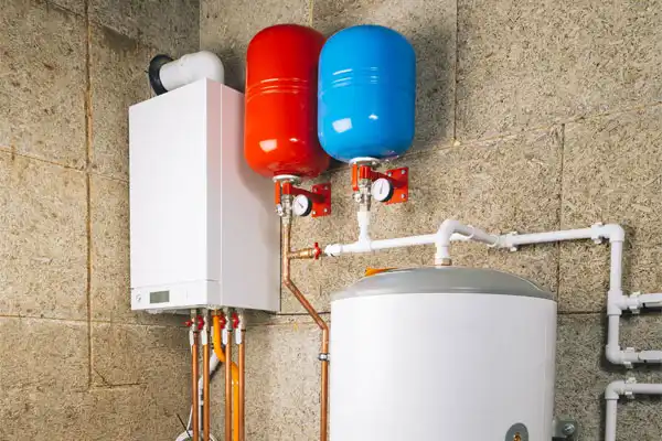 Heating system setup with a white boiler, red and blue expansion tanks, and connected pipes against a textured wall.