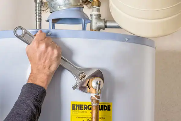 A person's hand uses a wrench to tighten a connection on a water heater with an EnergyGuide label.