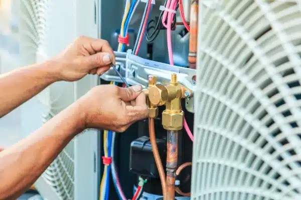 Hands of a technician using a wrench to adjust components on an HVAC unit, surrounded by various colored wires and metal parts.