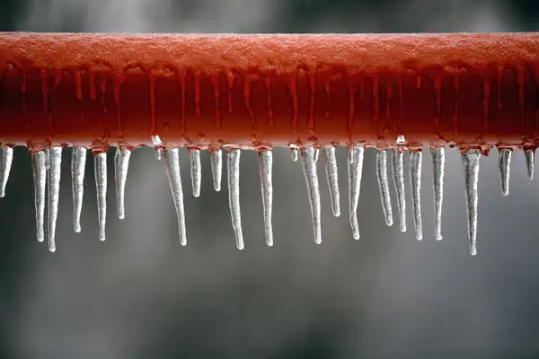 Icicles hanging from a red pipe against a blurred gray background.