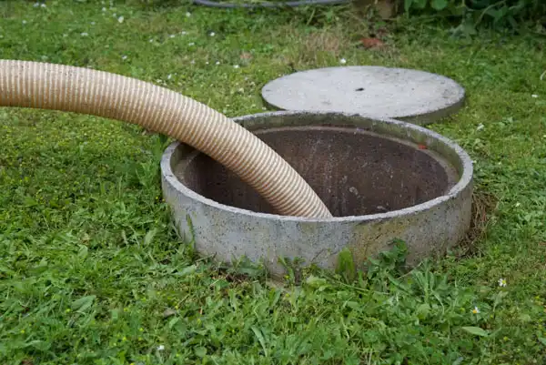 Hose inserted into an open septic tank on grass, with a removed circular concrete lid placed nearby.