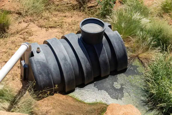 Black septic tank in dry, grassy area, partially submerged and connected to a white pipe.